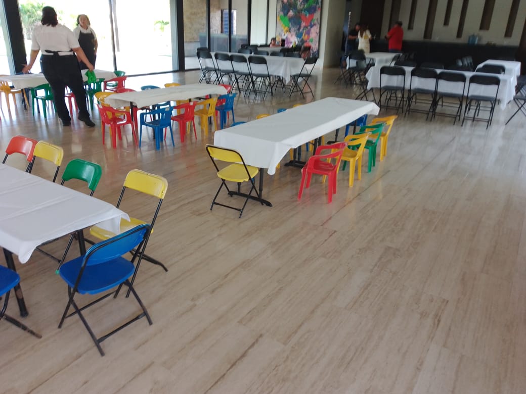 Mesas largas con mantel blanco y sillas infantiles de colores rojo, azul, amarillo y verde en un salón