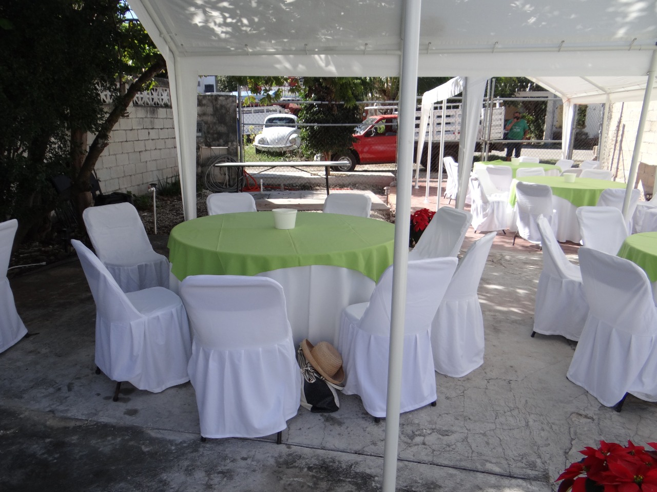 Mesas redondas con mantel blanco y cubremantel verde, rodeadas de sillas con funda blanca, montadas bajo una carpa