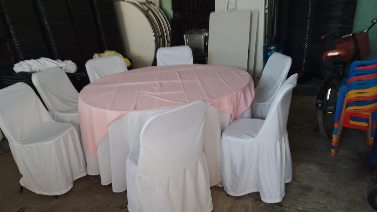 Mesa redonda con mantel blanco y cubremantel rosa, rodeada de sillas con funda blanca